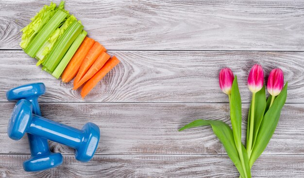 Весенняя спортивная композиция Flatlay с сельдереем и морковью голубыми гантелями и тюльпанами на сером деревянном фоне. Концепция здорового образа жизни, спорта и диеты весной. Вид сверху.