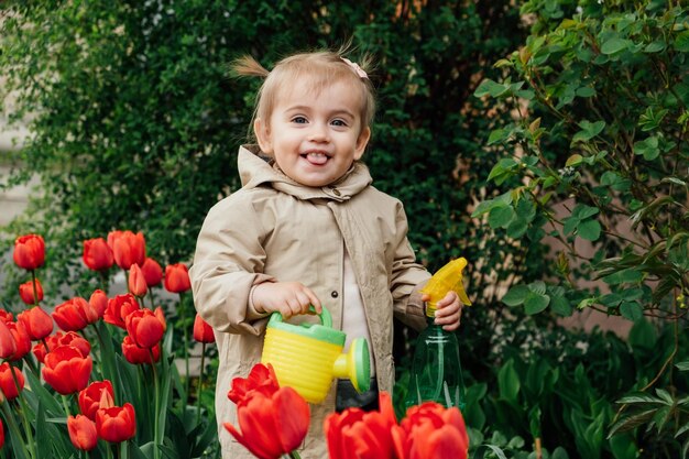 Фото Весеннее садоводство миленькая маленькая девочка в плаще поливает красные тюльпаны цветы в весеннем летнем саду