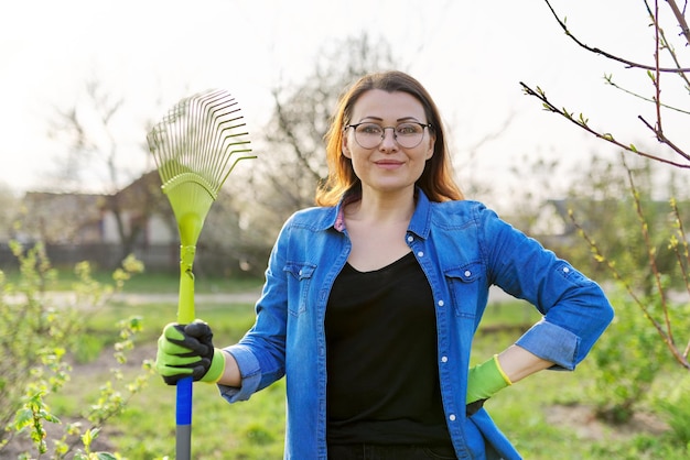 Spring gardening, portrait of mature smiling woman gardener with rake looking at camera. Trees and bushes background, seasonal cleansing of last year's grass and leaves in the garden, backyard