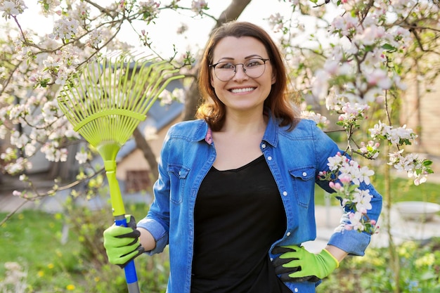 Spring gardening portrait of mature smiling woman with rake looking at camera Blooming trees in the garden background seasonal cleaning in the garden hobbies and leisure