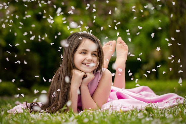 Spring girl Beautiful little girl in pink dress with smiling face lying barefoot on green grass in spring flower blossom petals outdoor