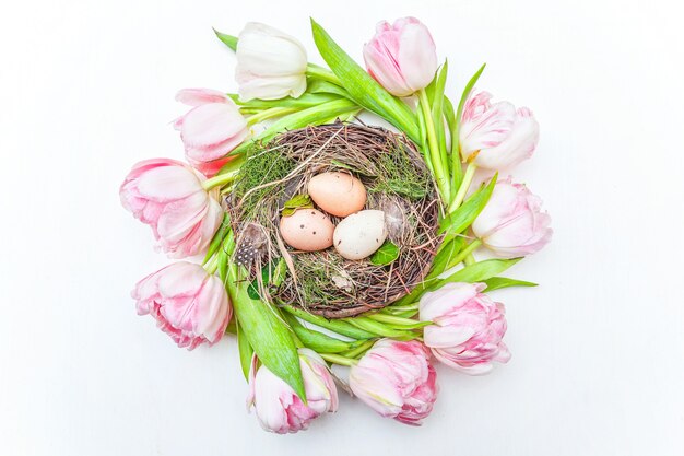 Spring greeting card. Easter eggs in nest and pink fresh tulip flowers bouquet on rustic white wooden background