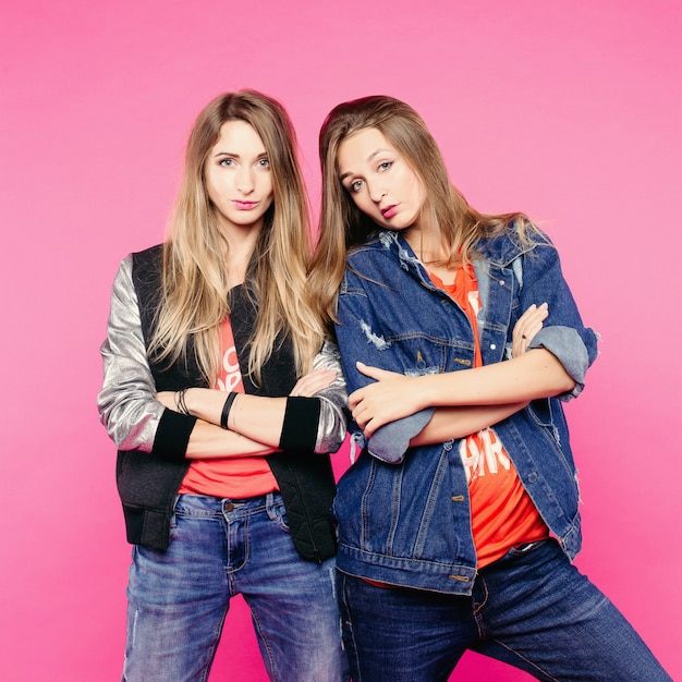 The spring image of two positive women with glasses, women friends with straight hair who hug. A woman a denim jacket puts a womanfriend's horns. In clothes of everyday style and glasses.