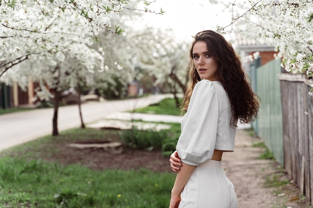 Spring lifestyle. Model posing near white blooming trees without mask outdoor countryside. Dreaming girl with curly hair.