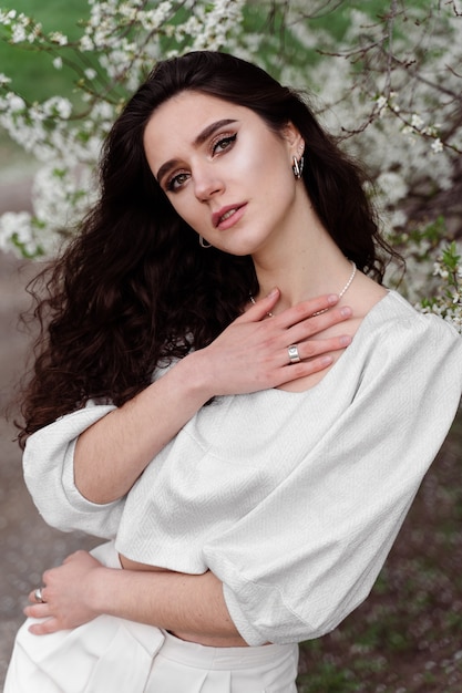 Spring lifestyle. Model posing near white blooming trees without mask outdoor countryside. Dreaming girl with curly hair.