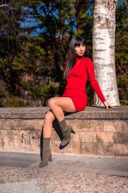 Spring lifestyle. Portrait of a young brunette in a park with a red dress resting and enjoying nature sitting next to a tree