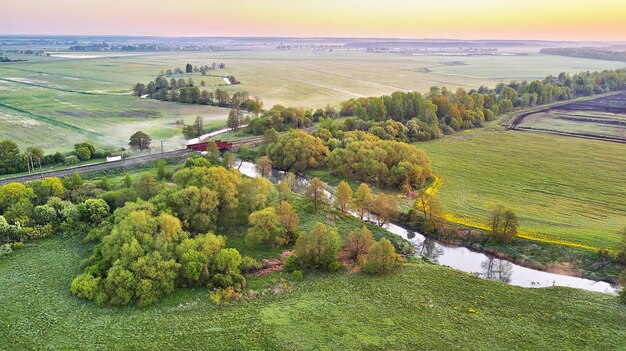 Фото Весенний утренний пейзаж железнодорожный мост пересекает спокойную реку на цветущих лугах вид с воздуха на сельскую местность сверху сельскохозяйственные желто-зеленые поля рапса сцена цветения одуванчика