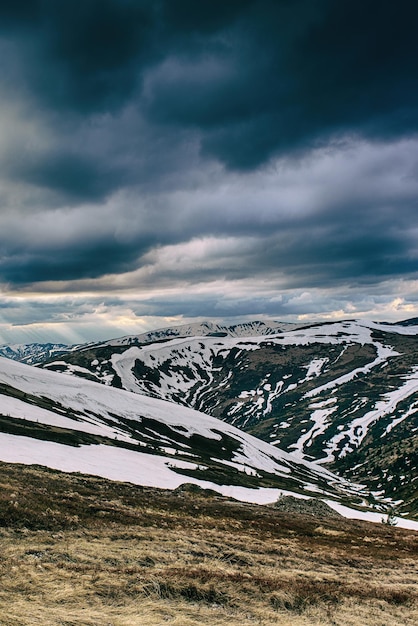 Весенний горный пейзаж со снегом и травой. Драматические облака лежат на горизонте и светит солнце. Естественный фон путешествия на открытом воздухе в стиле ретро-хипстер.