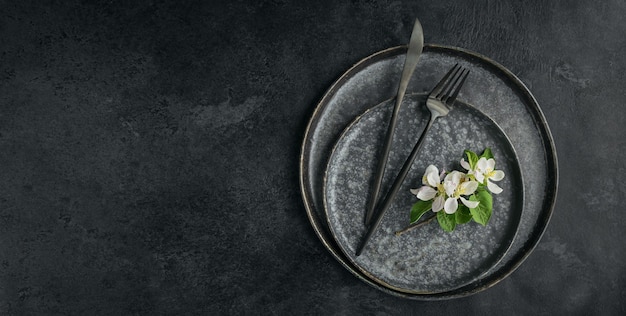 Spring place table setting with blossoming apple tree branches and flowers on black table. Holidays decoration on Provence style. Romantic dinner. Overhead with copy space for text