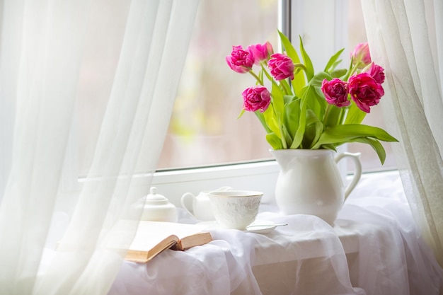 A spring still life a bouquet of tulips in a vase a cup of tea and an old book are lying on window