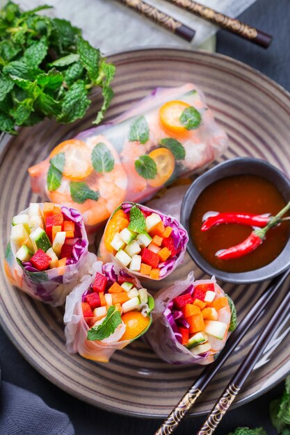 Spring or summer rolls with rice paper, carrot, chili sauce,
red cabbage, zucchini , pepper, shrimps and kumquats. tasty asian
food. top view flat lay background