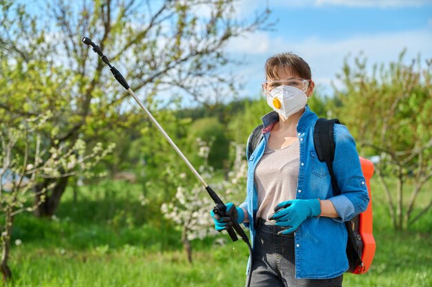 Spring summer work in garden backyard woman with goggles respirator backpack garden spray gun under pressure looking at camera Protection and care of plants from insect pests bacterial diseases
