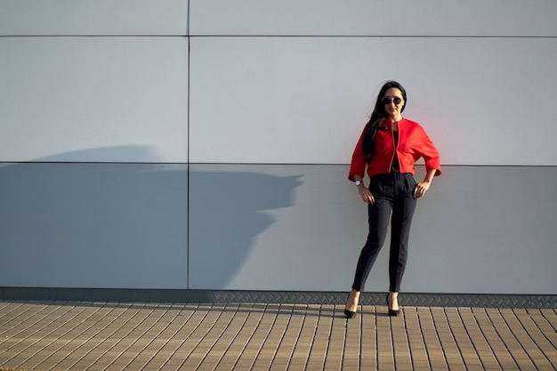 Spring trendy collection. Young attractive girl in fashionable sunglasses wearing stylish red jacket and blue pants on gray wall