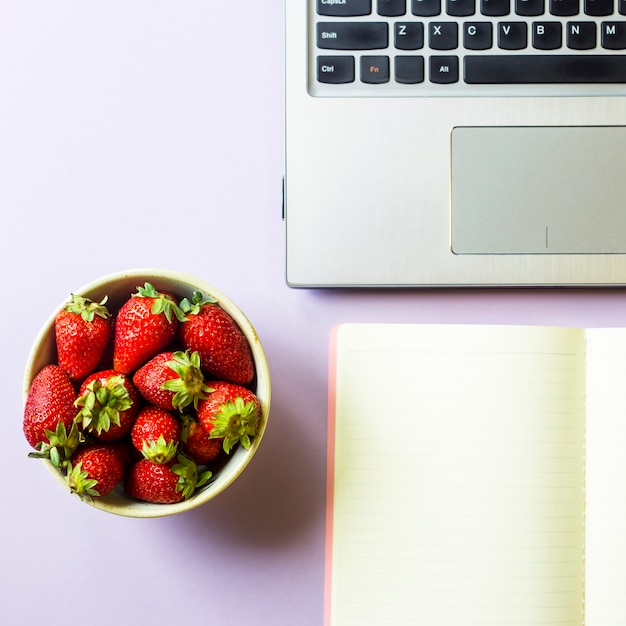 Spring workplace with laptop, notebook and a bowl of strawberries 