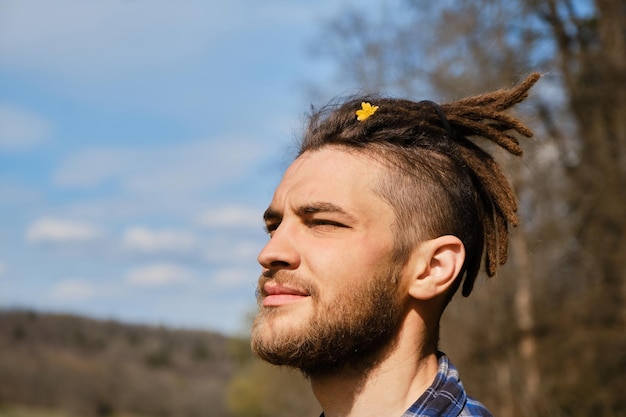 Spring yellow primrose anemone in mans hairstyle closeup portrait Close eyes from sun and pleasure Young Caucasian hipster with dreadlocks and beard in park on sunny summer day