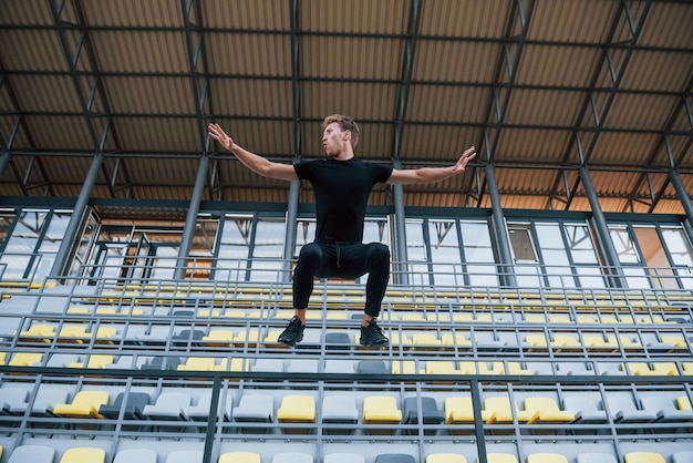 Springen en parkour doen op de tribunes Sportieve jonge kerel in zwart shirt en broek overdag buiten