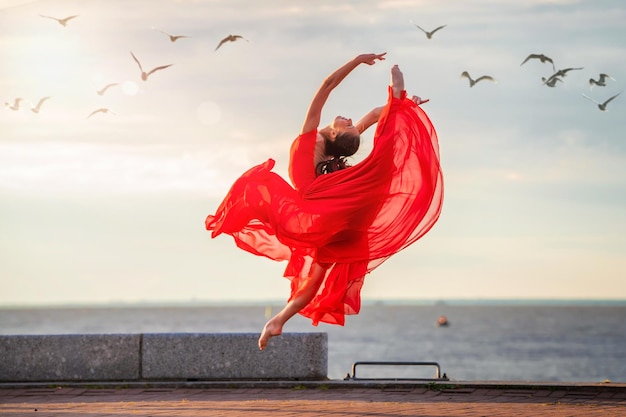 Springende ballerina in een rode vliegende rok en leotard op oceaandijk of zeestrand omringd door meeuwen in de lucht