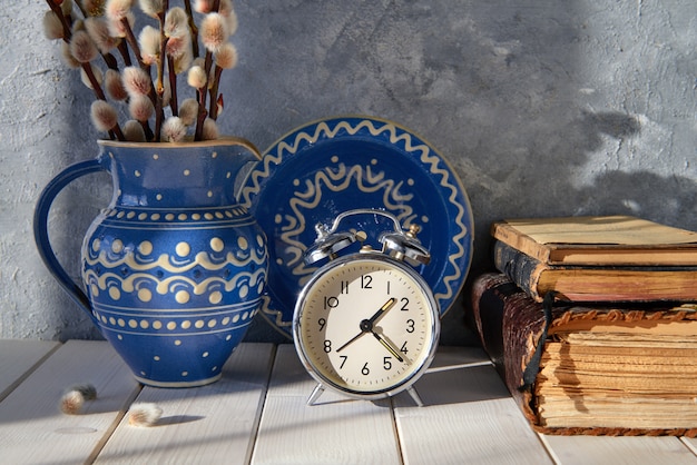 Springtime background with alarm clock, ceramic plate and a pitcher with pussy-willow and old books on wood