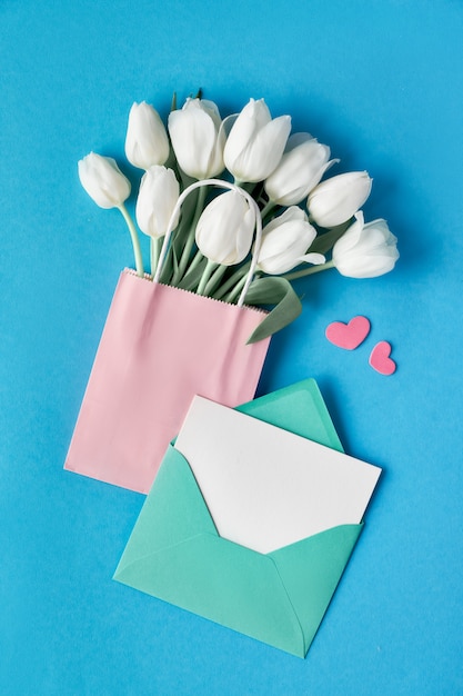 Springtime flat lay, white tulips in paper bag on blue mint background with envelope, blank card and decorative hearts