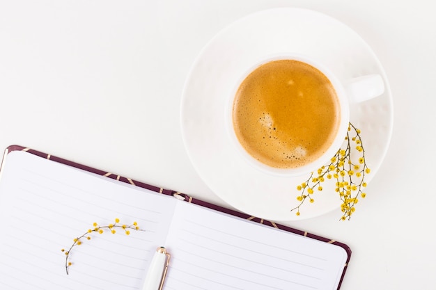 Luogo di lavoro di primavera con una tazza di caffè e un fiore di mimosa accanto a un blocco note aperto. vista dall'alto, piatta distesa