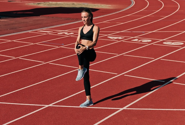 Sprinter woman in sportswear doing warm-up exercises before run on stadium track with red coating outdoor