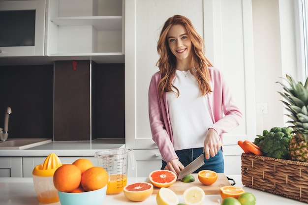 Sproeten gember vrouw lacht naar de camera tijdens het snijden van fruit en het maken van sap