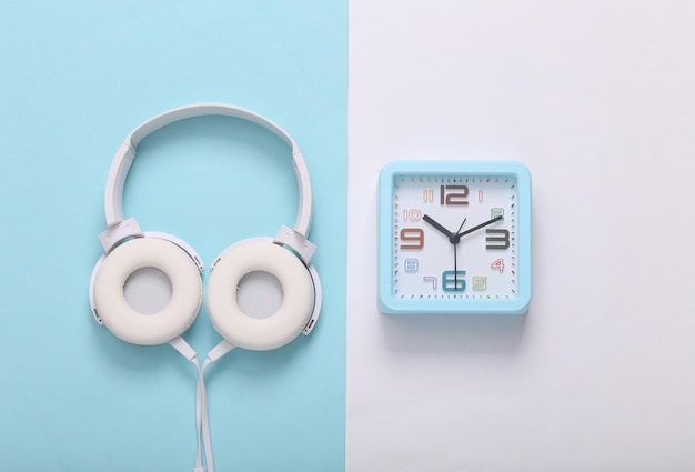 Square alarm clock with headphones on white blue background Music time Top view