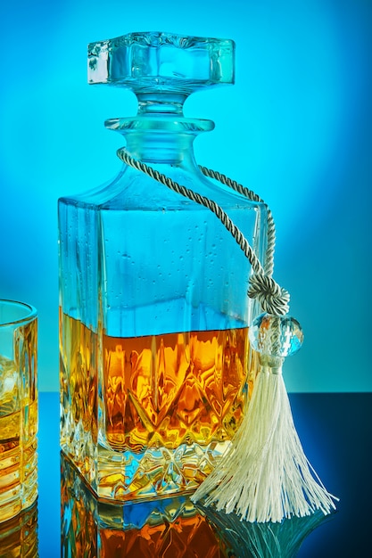 Square crystal decanter with scotch whiskey or brandy on a blue gradient background with reflection