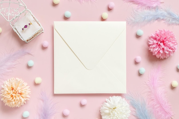 Квадратный конверт между помпонами пастельных цветов и перьями возле кольца в подарочной коробке на розовом