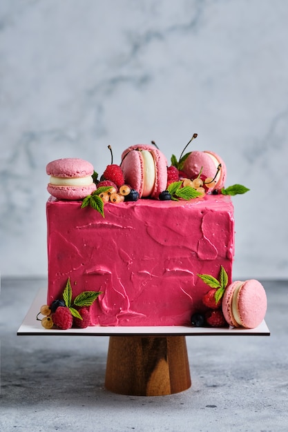 Square pink cake garnished with macarons and fresh berries. Cake for the holiday. Dessert is garnished with fresh raspberries, white currants, cherries and blueberries.