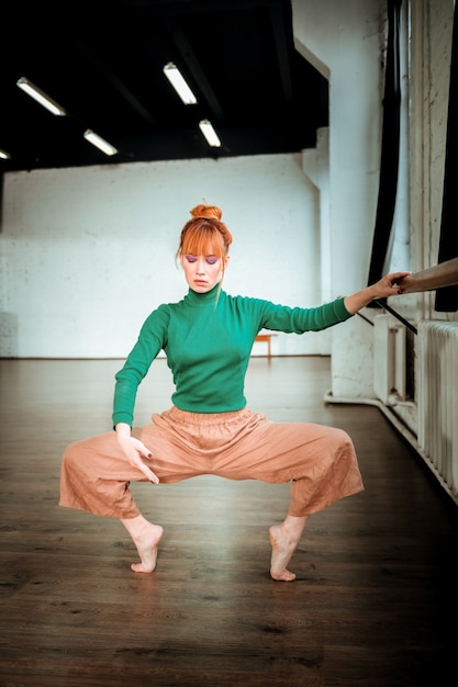 Squatting. Young professional modern dancer with red hair wearing a green turtleneck doing squatting