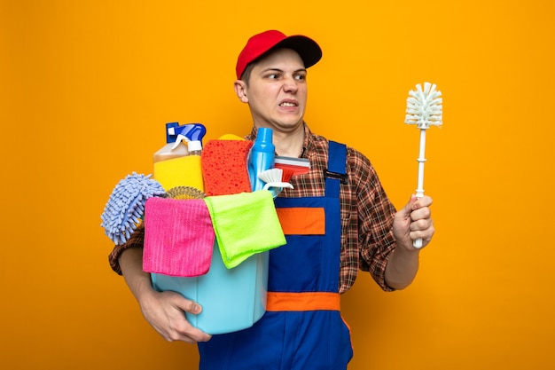 Photo squeamish young cleaning guy wearing uniform and cap holding bucket of cleaning tools and looking at brush in his hand
