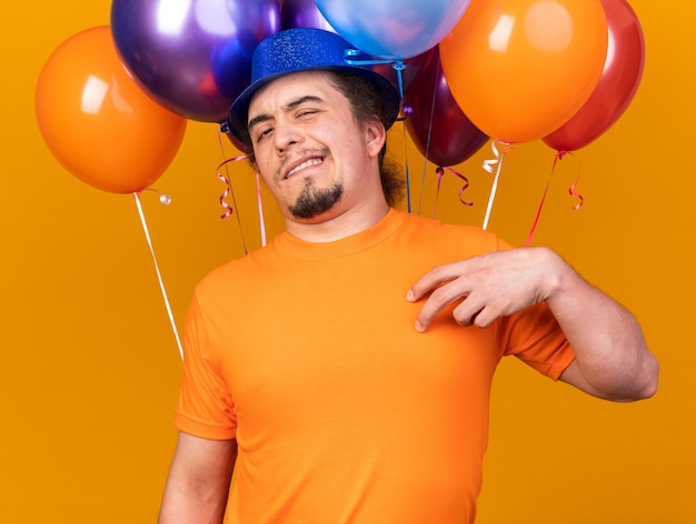 Squeamish young man wearing party hat standing in front balloons isolated on orange wall