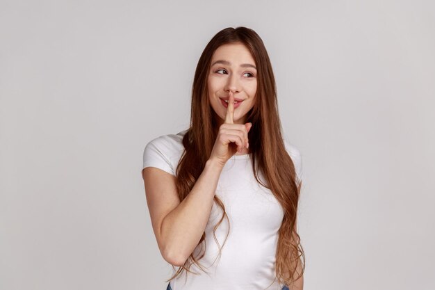 Sst, het is een groot geheim. Mooie sluwe vrouw die lacht, gebaar geheim teken toont met vinger in de buurt van haar lippen, wit T-shirt dragend. Indoor studio opname geïsoleerd op een grijze achtergrond.