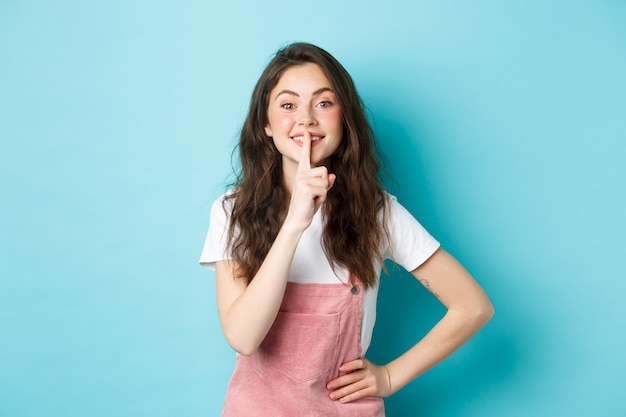 Sst stil. schattige brunette meisje zwijgt en glimlacht, heeft een geheim, houdt de vinger op de mond en kijkt vriendelijk naar de camera, staande over een blauwe achtergrond