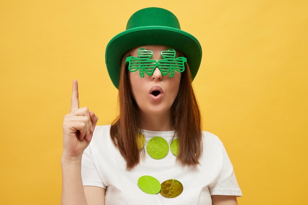 St Patrick's Day festiviteiten Ierse hoed en klaverviering Geïnspireerde vrouw met feestelijke groene hoed en shamrock bril staat geïsoleerd op een gele achtergrond met de vinger omhoog met een idee