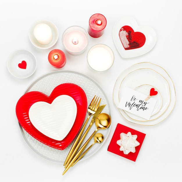 St. Valentine Day table setting. White and red dishes in form of heart, candles and cutlery white background. Romantic concept