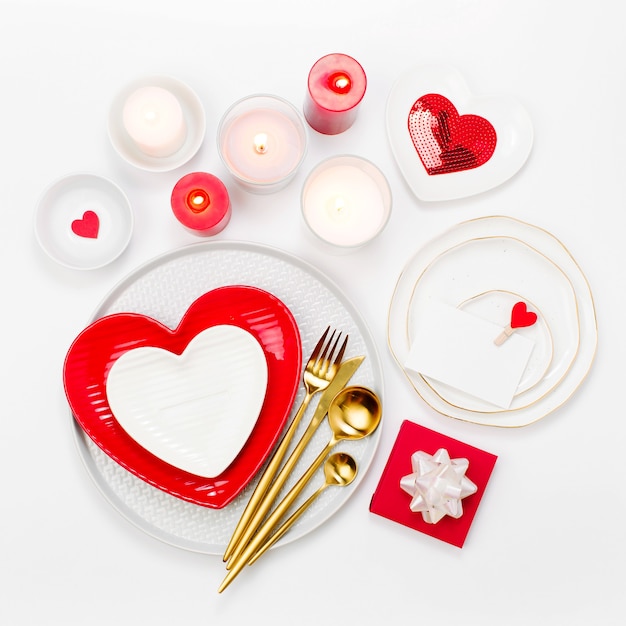 聖バレンタインデーのテーブルセッティング。ハート、キャンドル、カトラリーの白い背景の形で白と赤の料理。ロマンチックなコンセプト
