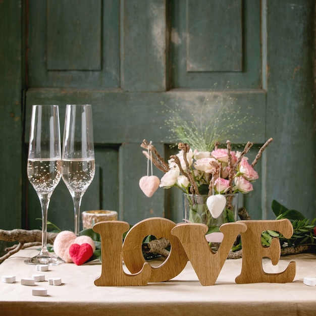 St. Valentines day or wedding romantic table setting with wooden letters love, needle felted hearts, bouquet of pink roses, couple glasses of champagne on natural linen tablecloth. Square image