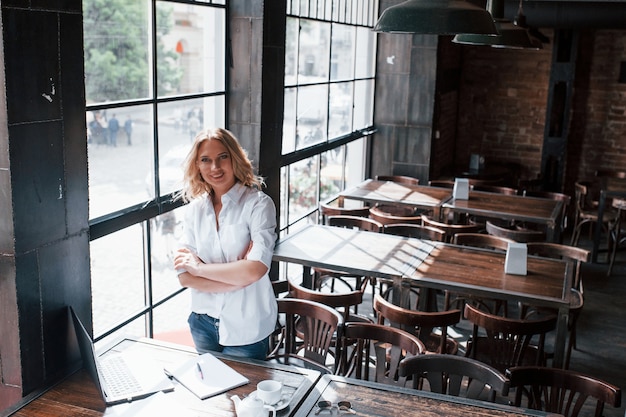 Staand met gekruiste armen. Zakenvrouw met krullend blond haar binnenshuis in café overdag.