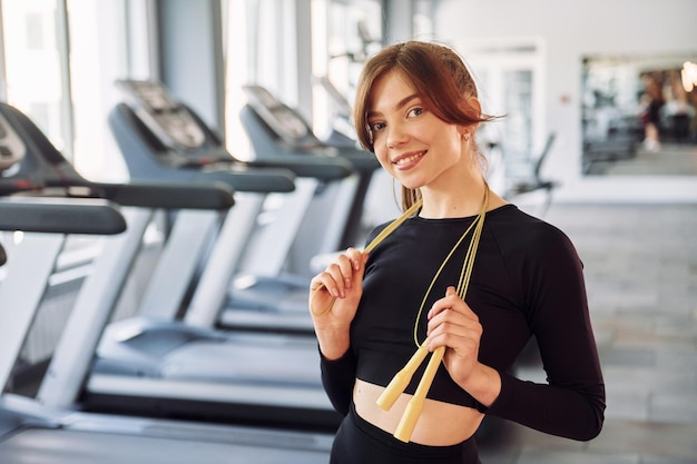 Staande met springtouw en glimlachend Jonge mooie vrouw in sportieve kleding heeft een fitnessdag in de sportschool