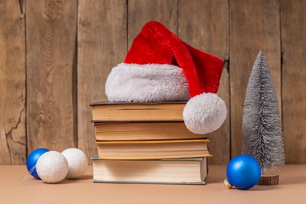 Pila di libri, decorazioni natalizie e cappello di babbo natale su fondo di legno.