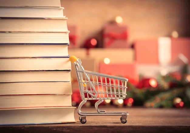 Stack of books and shopping cart with Christmas gifts on background