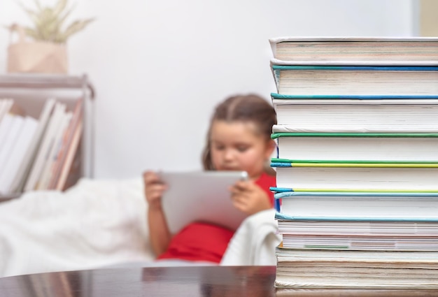 A stack of books on the table against the background of a child in a chair playing a tablet