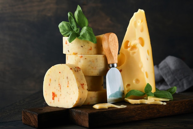 暗い木製のテーブル背景にサービング ボードにトマトと調味料とスイス チーズとバジルとナイフのチーズのスタック さまざまなチーズの種類の品揃え チーズの背景