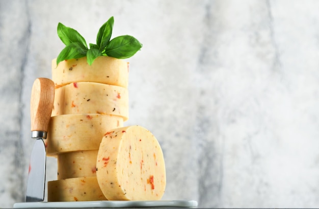 Stack of cheese with tomatoes and seasoning and Swiss cheese on with basil and knife on serving board on light grey table background Assortment of different cheese types Cheese background