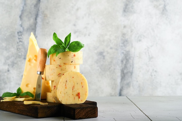 Stack of cheese with tomatoes and seasoning and Swiss cheese on with basil and knife on serving board on light grey table background Assortment of different cheese types Cheese background