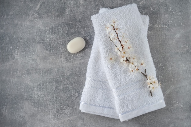 Stack of clean soft towels with blooming cherry branches and soap  on light gray background. SPA concept. Minimalism, soft focus, top view.