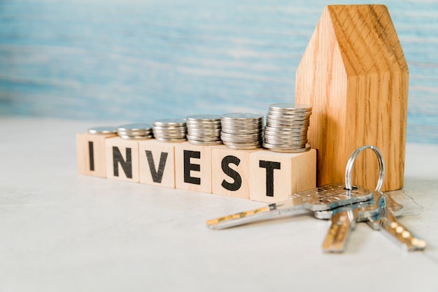 Photo stack of coins over the invest wooden blocks near the house model with silver keys on white surface