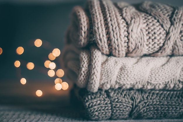 Photo stack of cozy knitted sweaters and garland lights on wooden background.